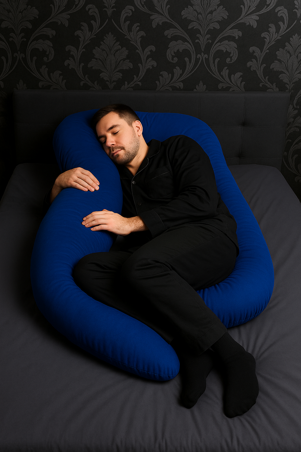 Man lying on a blue HerPillow body pillow U-shaped pillow against a dark patterned wall.